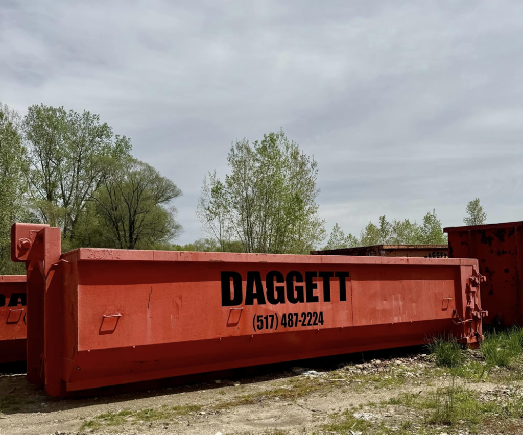 An orange dumpster with 'DAGGETT' branding from Daggett Container Service LLC, placed in a grassy area in Lansing, MI.