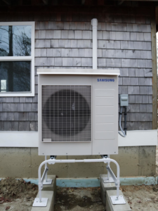 An outdoor Samsung heat pump unit installed on a sturdy stand next to a shingle-sided house by Coastal Home Improvements in Greenwood, DE.
