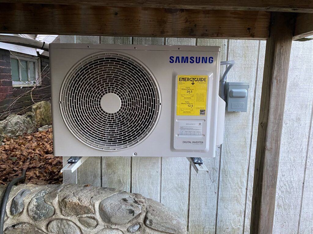An outdoor Samsung heat pump unit with an EnergyGuide label, mounted under a wooden deck by Coastal Home Improvements in Greenwood, DE.