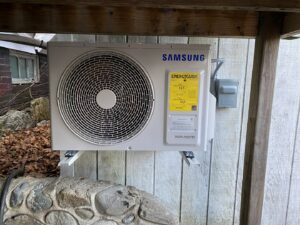 An outdoor Samsung heat pump unit with an EnergyGuide label, mounted under a wooden deck by Coastal Home Improvements in Greenwood, DE.