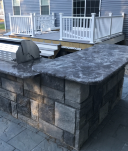 An outdoor kitchen island with a built-in grill and custom countertop, next to a deck, by Burton Restoration LLC in Bend, OR.