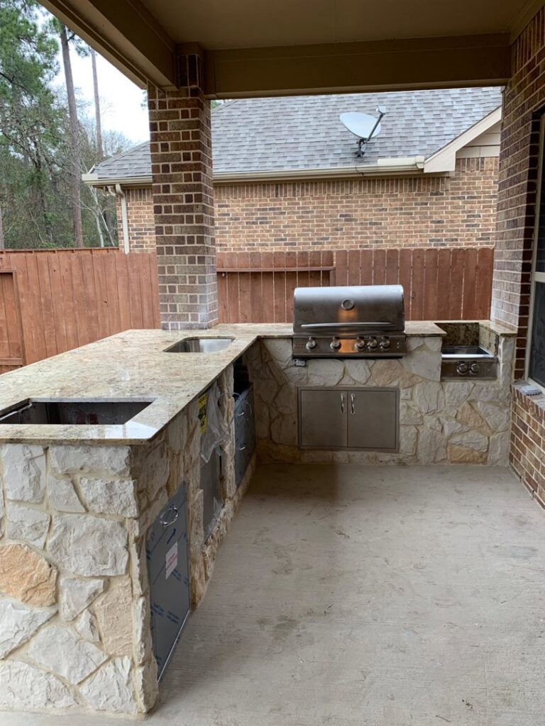 An outdoor kitchen under a covered patio with stone veneer, granite countertops, and a built-in grill by MC Master Construction in League City, TX