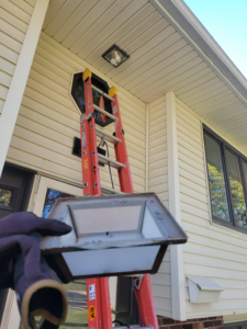A gloved hand holding an outdoor light fixture cover, with a ladder set up for replacement or repair by Cedar Valley Maintenance and Handyman Services LLC in Waterloo, IA.
