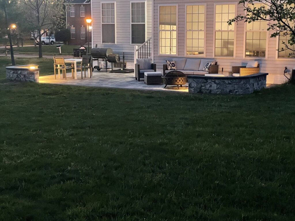 A newly installed outdoor paver patio with a fire pit and stone retaining walls, lit up at night by Landscape By Today in Bowie, MD.