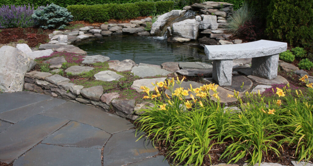 An outdoor landscape featuring a stone pond, waterfall, and patio with a stone bench by Adam Ross Cut Stone in Albany, NY.