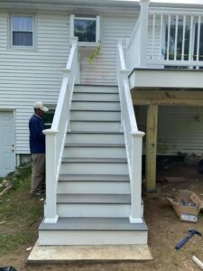 A worker standing next to a newly installed outdoor staircase with white railings and gray steps, completed by Men Construction in Lowell, MA.
