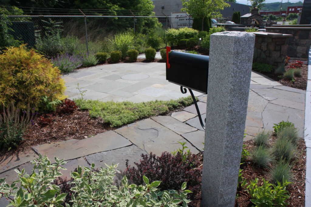 An outdoor stone patio with a granite mailbox post and lush landscaping by Adam Ross Cut Stone in Albany, NY.