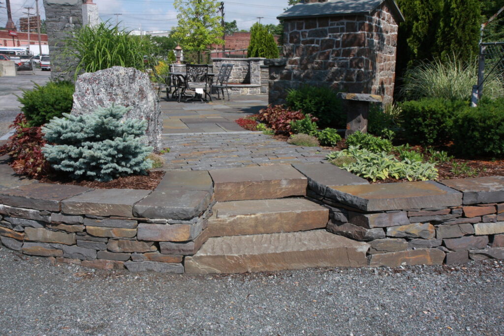 Outdoor stone steps leading to a patio area with a stacked stone retaining wall by Adam Ross Cut Stone in Albany, NY.