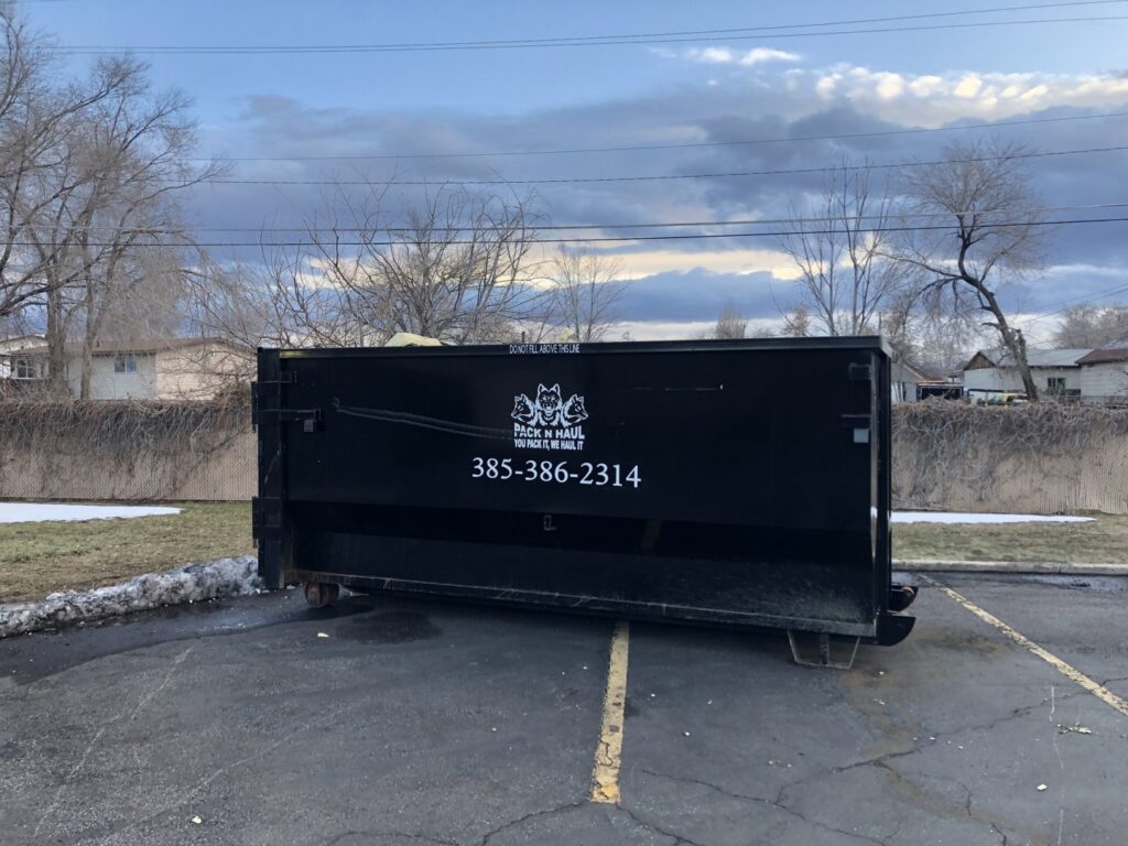 A large black dumpster with the Pack N Haul logo and contact information, placed in a parking lot in West Valley City, UT.
