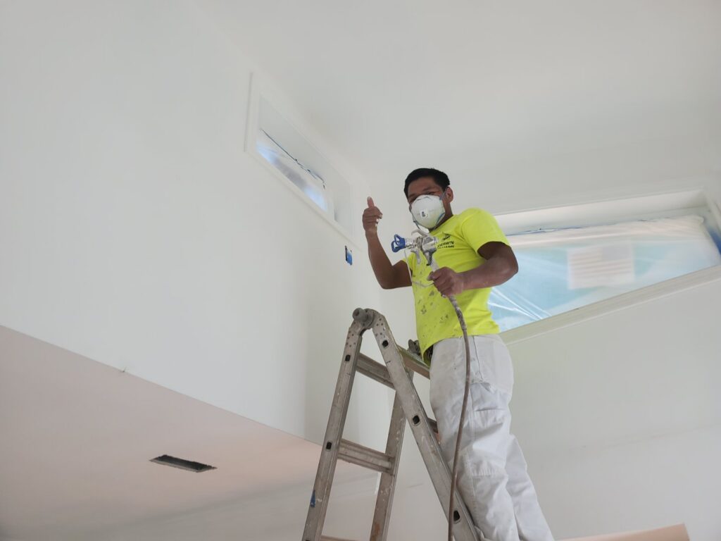 A painter wearing a mask and holding a spray gun on a ladder, giving a thumbs up, during a painting job by GCI Services, Inc. in Simpsonville, SC