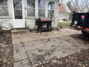 An outdoor patio with a grill and scattered debris, with a junk removal trailer from Rosales Junk Removal in Des Moines, IA.