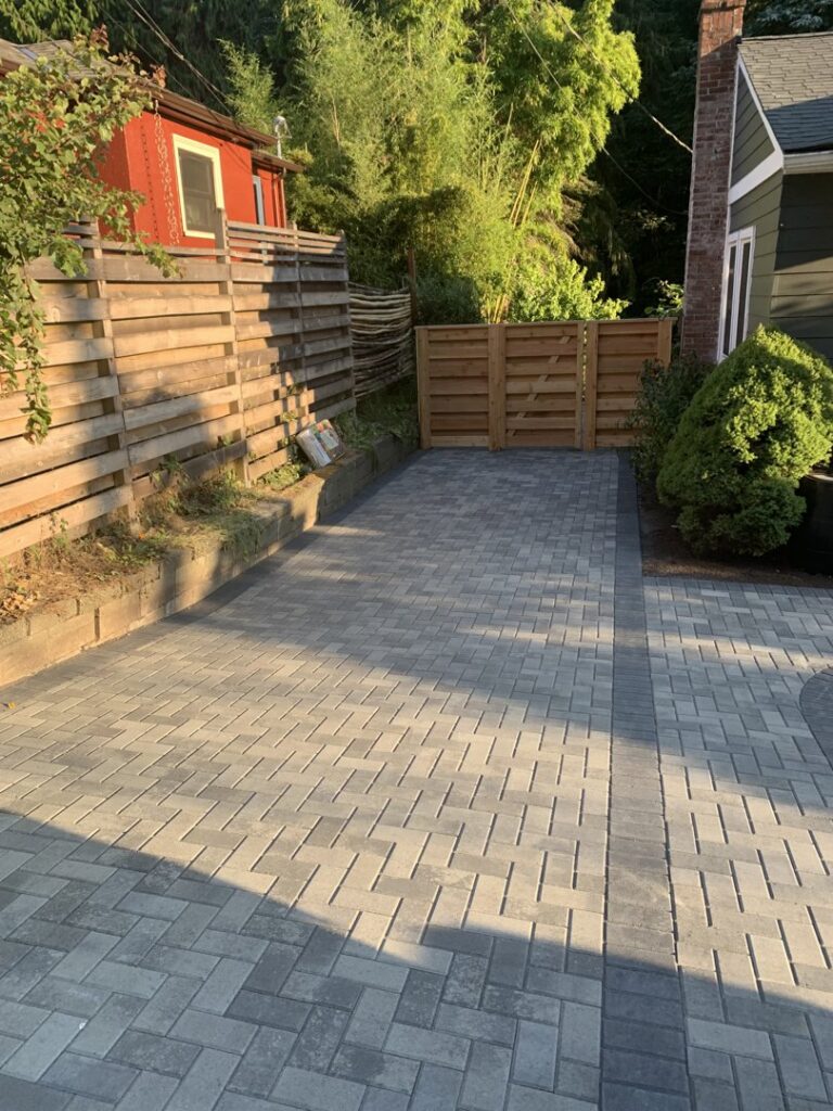 A paved driveway or patio area with a wooden fence and gate in the background by LMS Landscapes & Fence services in Auburn, WA.