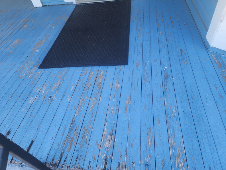 A wooden porch floor with peeling blue paint and a black doormat, showing a repair need for P V A Construction Managers in Albany, GA.