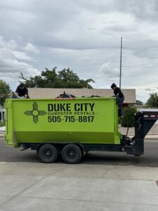 Two individuals actively loading various junk items into a bright green Duke City Dumpster Rentals LLC dumpster on a trailer in Albuquerque, NM.