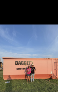 Two people standing with a pink Daggett Container Service LLC dumpster, ready for junk removal in Lansing, MI.