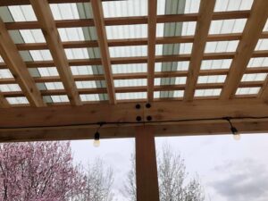 Underside of a newly constructed pergola roof with wooden beams, corrugated panels, and string lights by Fisher Construction, LLC in Selah, WA.