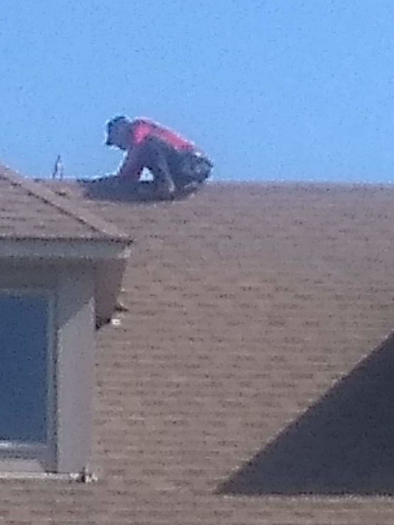 A person crouching on a brown shingled roof, performing work for Superior Roof Support in Milano, TX.