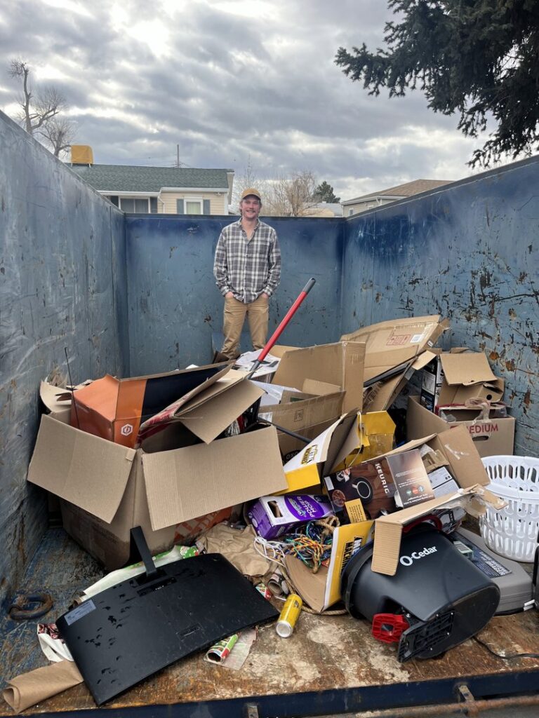 A person standing inside a blue dumpster, loading it with cardboard boxes and household junk for Blue Bin Dumpster Rentals in Salt Lake City, UT.