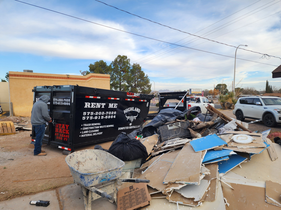 A person standing next to a Highland Dumpster Rentals LLC dumpster with a large pile of debris in Las Cruces, NM.