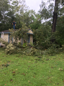 A person on a roof clearing storm debris and fallen branches from a small house, a service provided by H&H Construction of Columbus, LLC in Columbus, GA.
