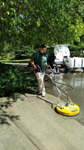 A person using a surface cleaner to pressure wash a concrete driveway, with a work van in the background, by JAMS Property Preservation LLC in Dayton, OH.