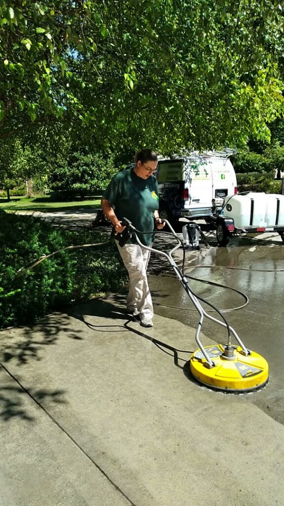 A person using a surface cleaner to pressure wash a concrete driveway, with a work van in the background, by JAMS Property Preservation LLC in Dayton, OH.