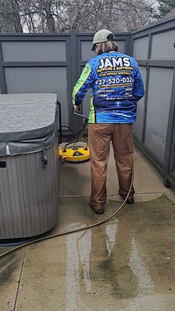 A person in a JAMS branded shirt using a surface cleaner to pressure wash a concrete patio next to a hot tub in Dayton, OH.