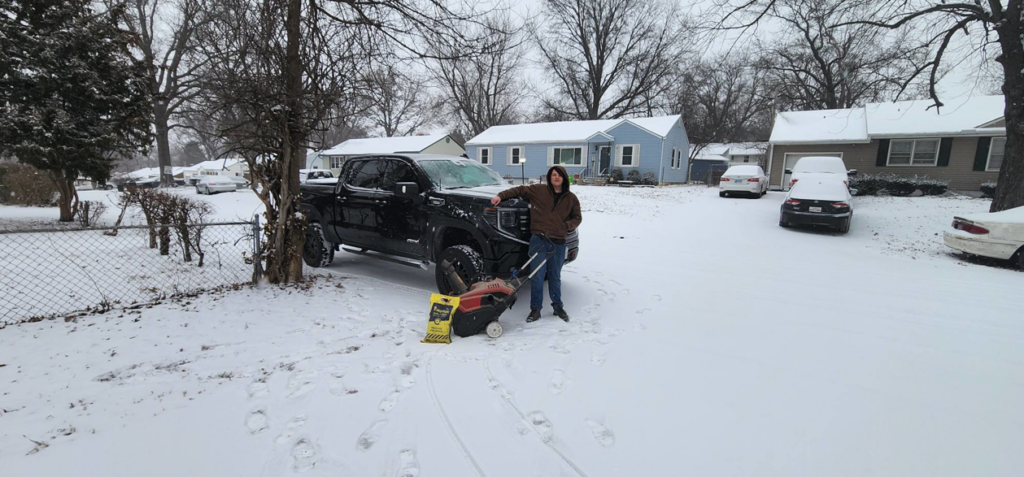 A person standing with a snow spreader and pickup truck, ready for snow removal services by Brother's Keeper Construction in Spokane, WA