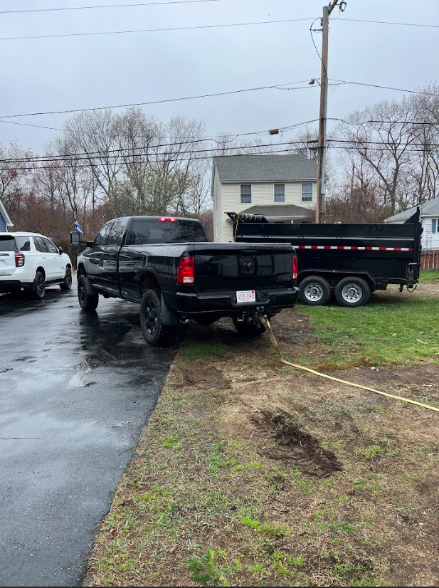 A black pickup truck pulling a dump trailer, parked in a residential area by Getting Trashed Dumpster Rentals & Excavation in Assonet, MA.