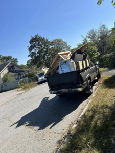 A pickup truck with a dump bed extension loaded with bulky junk and debris on a residential street by Trash Hogs in Worcester, MA.
