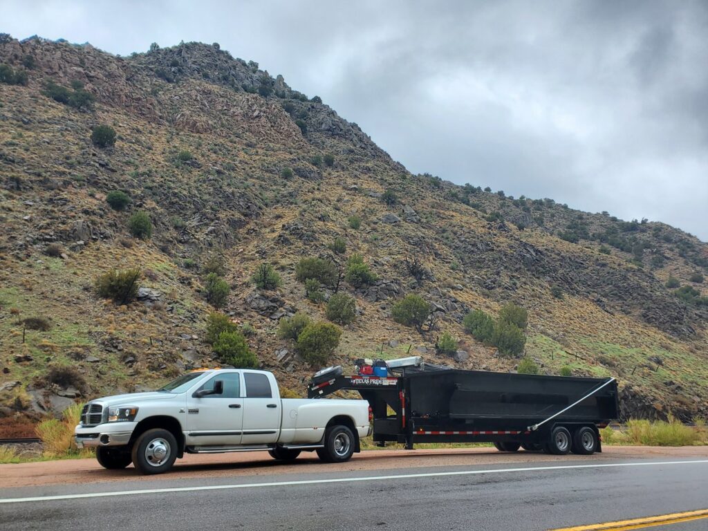 A white pickup truck towing a black dump trailer on a mountain road for Arkansas Valley Roll-Off in Salida, CO.
