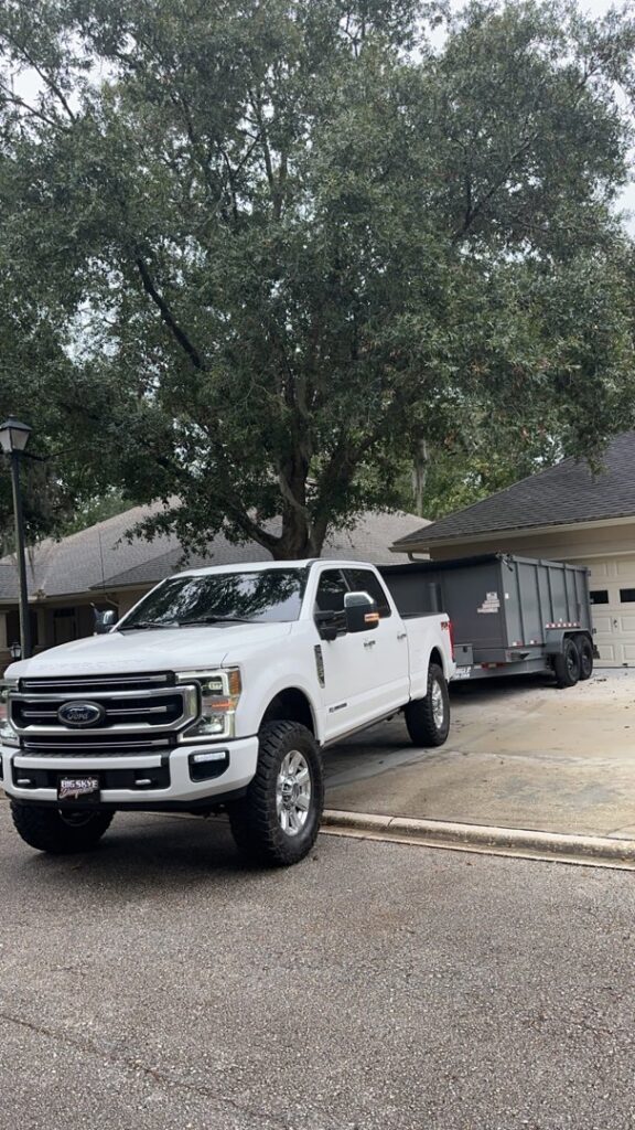 A white pickup truck towing a gray dumpster trailer from Big Skye Dumpsters on a residential street in Jacksonville, FL.