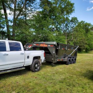 A white pickup truck towing a large green dumpster trailer in a grassy area, used by HD Waste Services for junk removal in Chattanooga, TN.