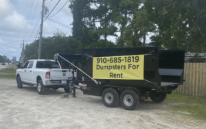 A white pickup truck towing a black dumpster trailer with a 'Dumpsters For Rent' banner for The Dumpster Dad in Wilmington, NC.