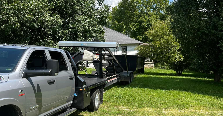 A grey pickup truck with a flatbed trailer carrying a roll-off dumpster for O'Lakes Dumpster Rental in Lexington, KY.