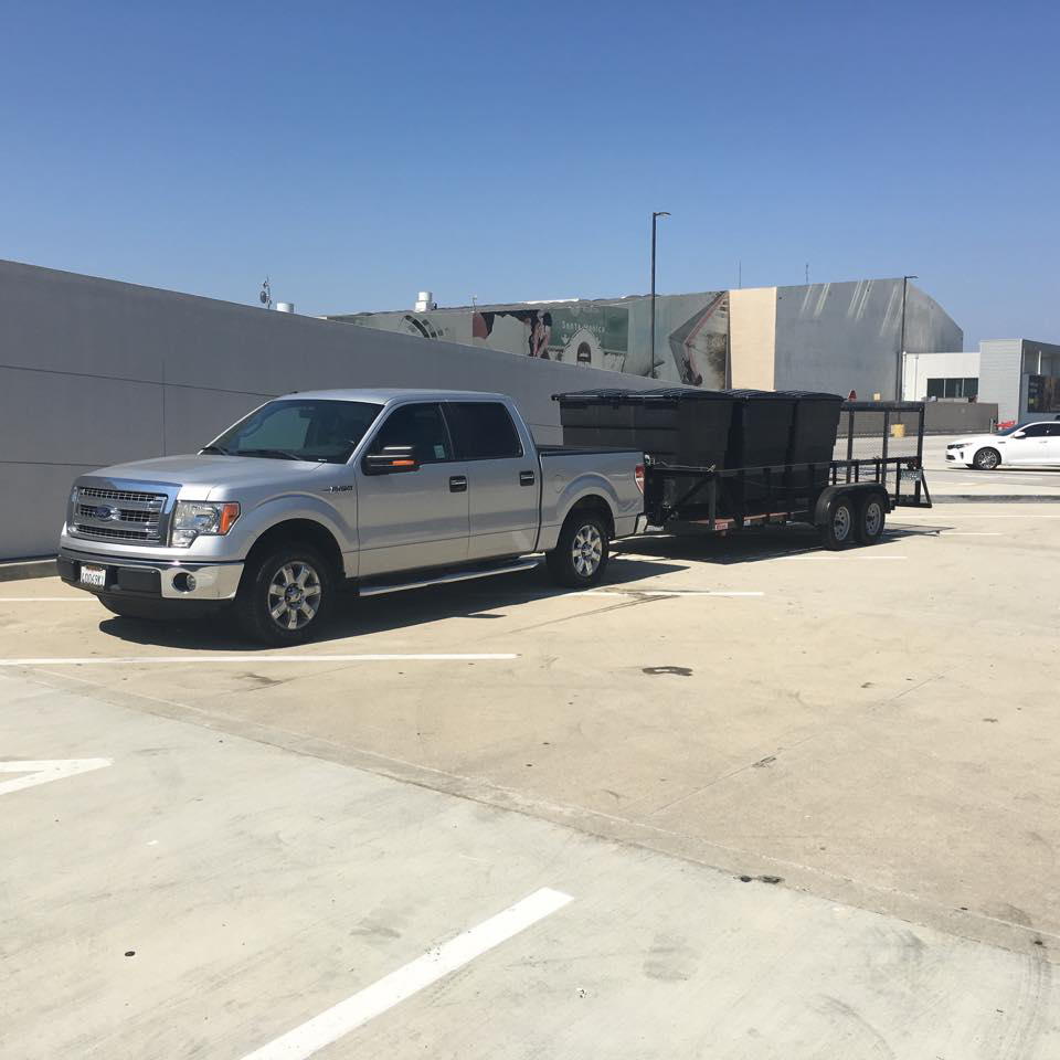 A silver pickup truck with a trailer loaded with black junk removal bins for Industry Services, LLC in Inglewood, CA.