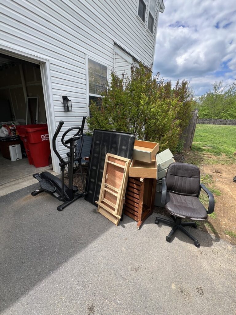 A pile of household junk including an elliptical, table, and chair outside a garage, ready for removal by Dumpin' Junk in Martinsburg, WV.
