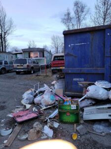 A large pile of household junk, bags, and debris next to a blue dumpster, for junk removal by Alaska Waste in Anchorage, AK.