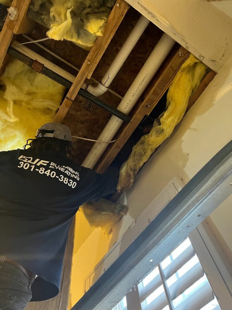 A plumber from JF Plumbing & Heating working on pipes and insulation in a ceiling space in Rockville, MD.