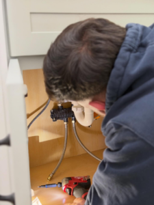 A plumber working on pipes under a sink with a power tool for Sandobros Plumbing in Charleston, SC