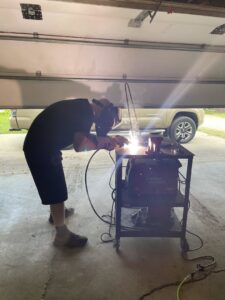 A professional handyman wearing a welding mask and gloves, actively performing welding work in a garage for Cedar Valley Maintenance and Handyman Services LLC in Waterloo, IA.