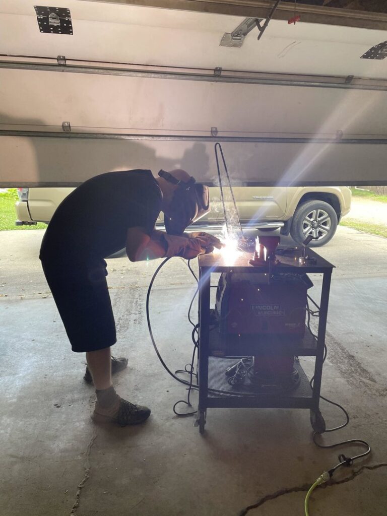 A professional handyman wearing a welding mask and gloves, actively performing welding work in a garage for Cedar Valley Maintenance and Handyman Services LLC in Waterloo, IA.