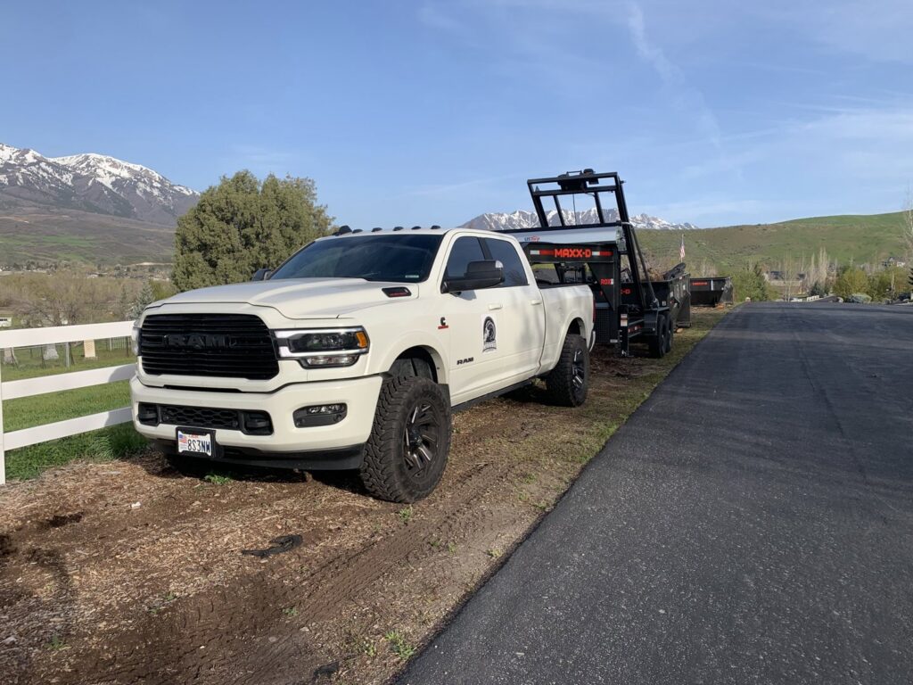 A RAAAM Dumpster Rentals truck pulling an empty roll-off dumpster trailer with a mountain view in Salt Lake City, UT.