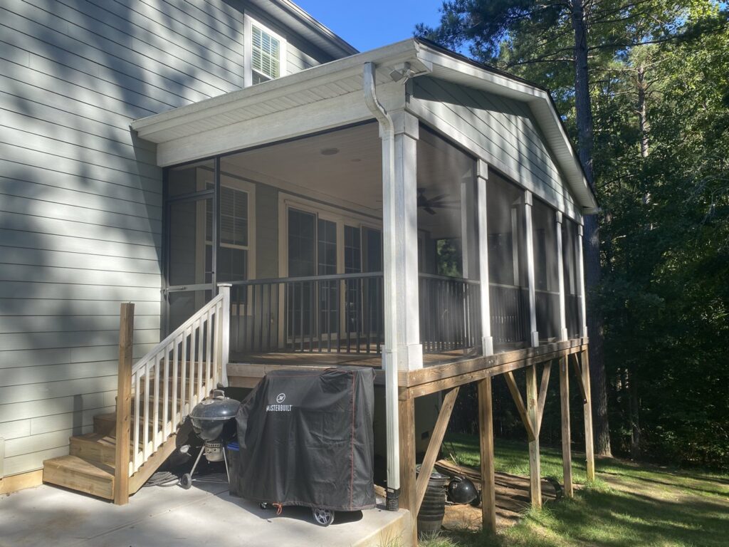 A raised screened porch with wooden stairs leading down to a patio with a grill, built by Screen Enc in Denver, NC