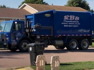 An RBS Sanitation Inc. blue garbage truck on a residential street, collecting waste from a bin in Tea, SD.
