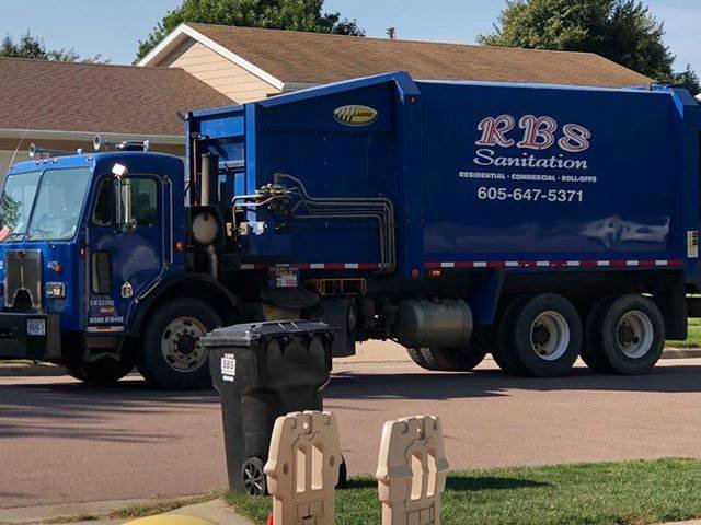 An RBS Sanitation Inc. blue garbage truck on a residential street, collecting waste from a bin in Tea, SD.