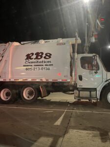 A white RBS Sanitation Inc. garbage truck on a street at night, performing waste collection services in Tea, SD.