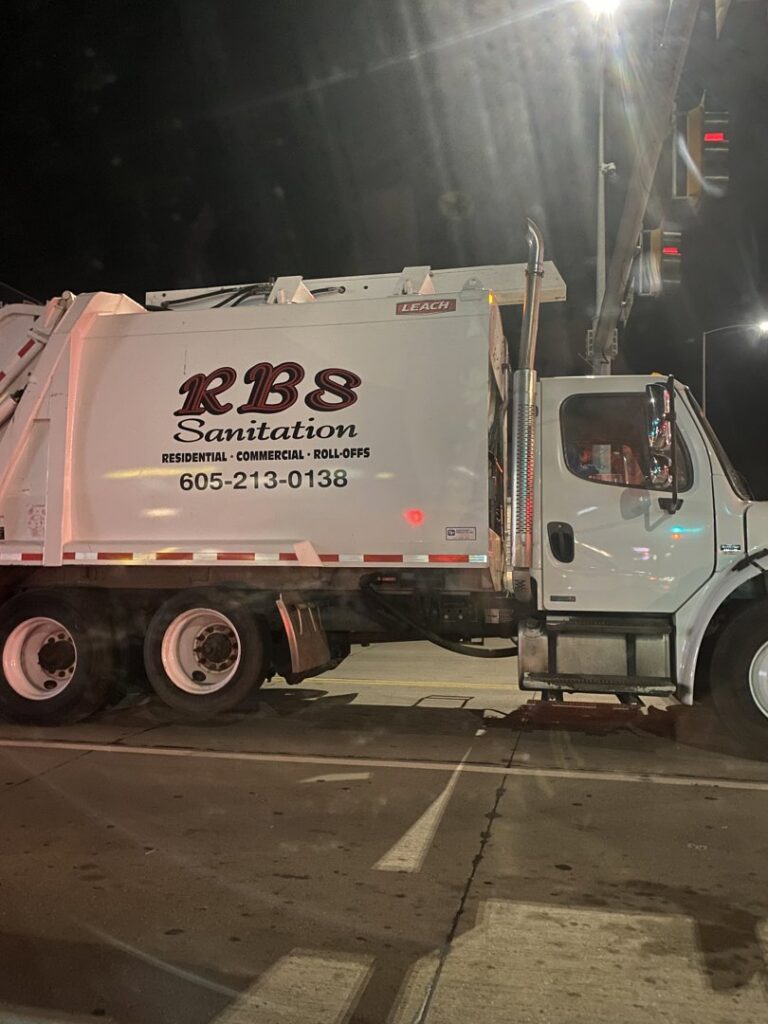 A white RBS Sanitation Inc. garbage truck on a street at night, performing waste collection services in Tea, SD.