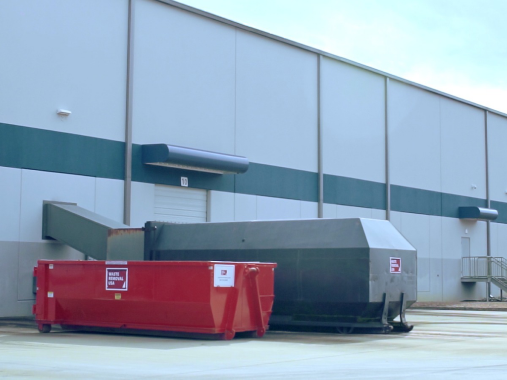 A red roll-off dumpster from Waste Removal USA placed on the ground next to an industrial compactor in Austin, TX