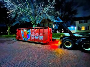A red roll-off dumpster from AT Disposal being picked up by a truck at night in Dewitt, IA.